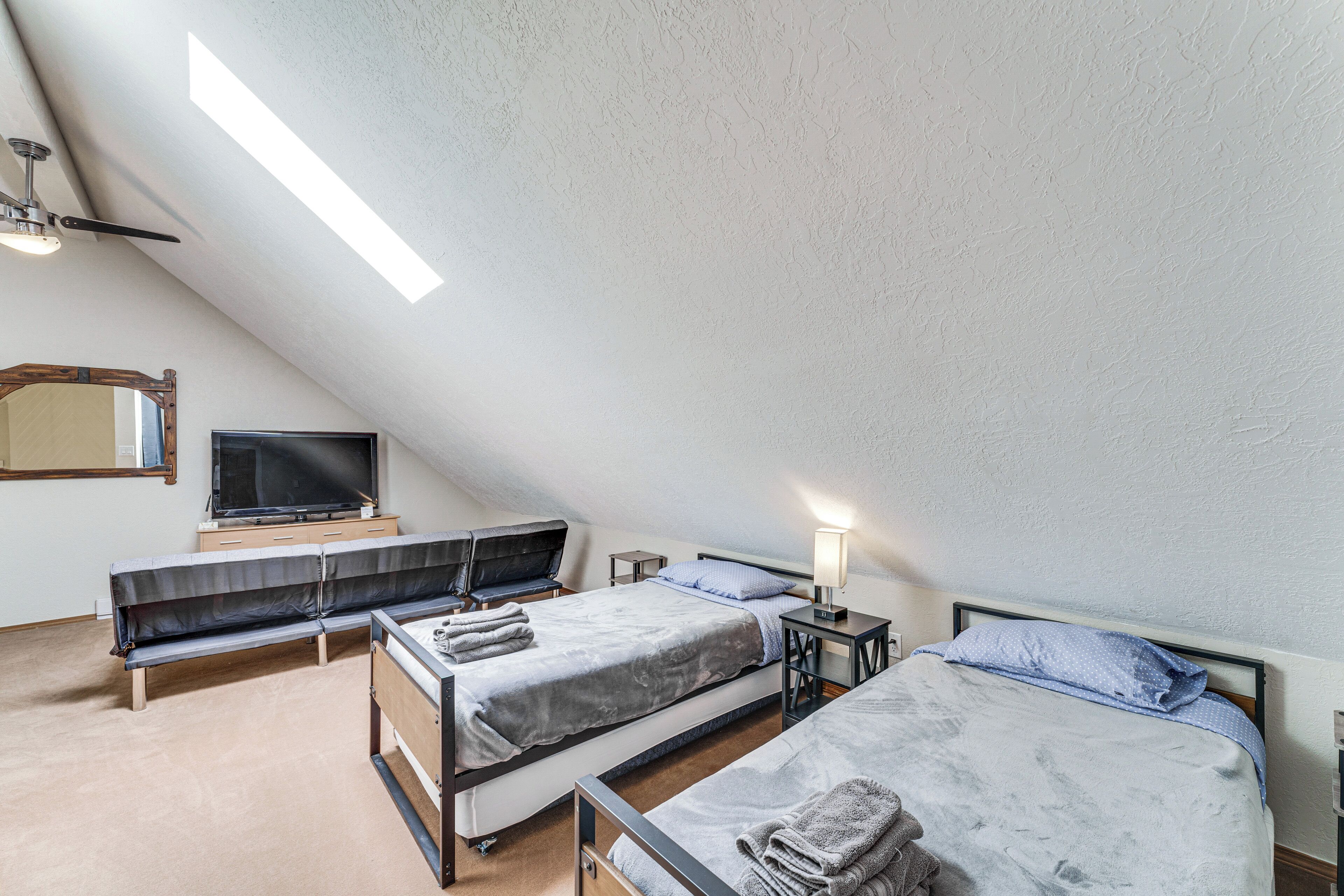 Quarto pequeno e bem iluminado com duas camas, uma televisão e um banco.