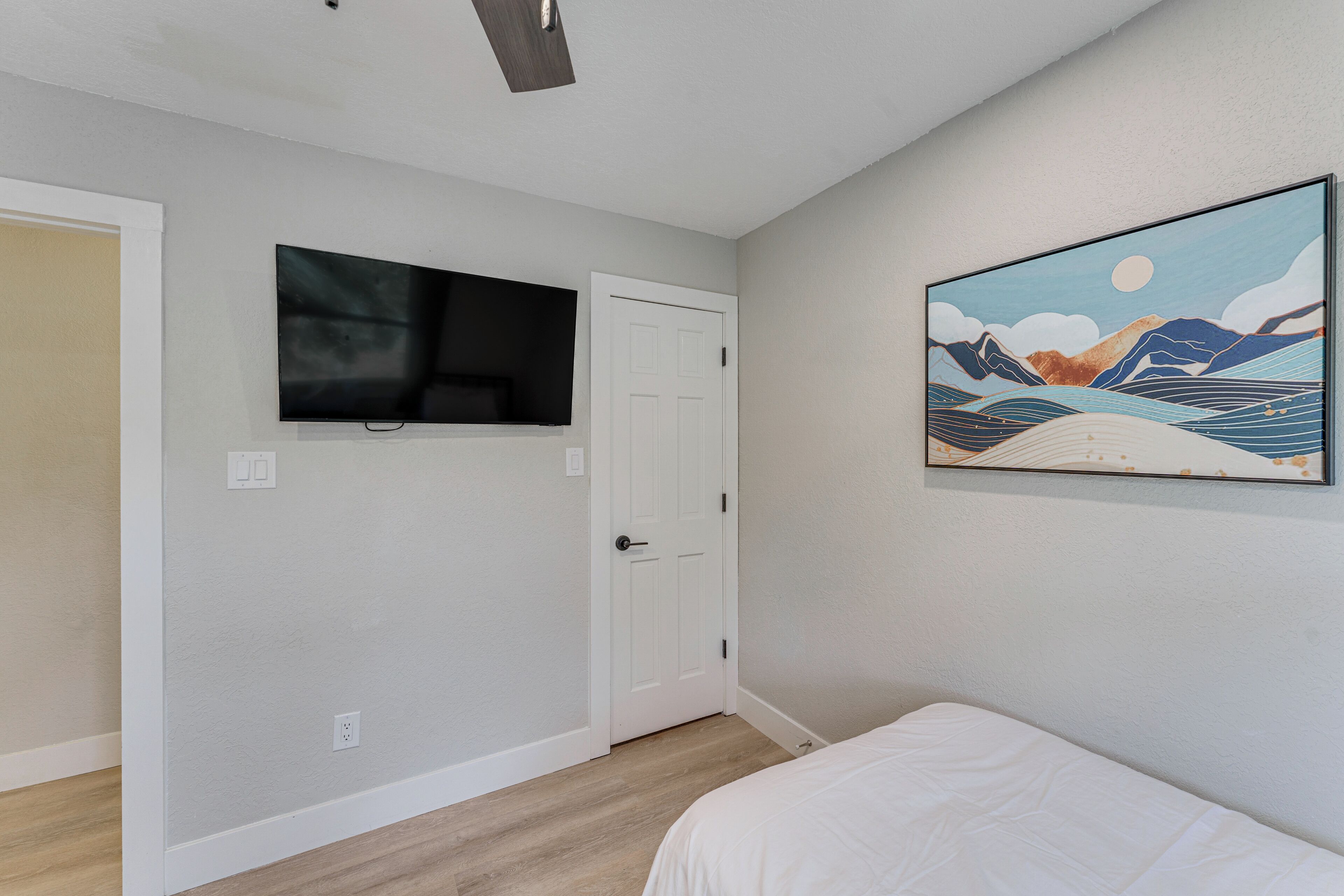 A bedroom with a bed, a mounted TV, a door, and a wall-mounted painting.