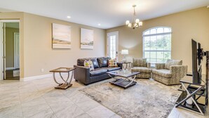 A spacious living room with a dark leather sofa, a glass coffee table, and a patterned armchair. There is a large window providing natural light and a chandelier hanging from the ceiling. - 5 Mi to Weeki Wachee Springs! Poolside Retreat (Brooksville)