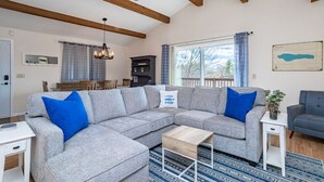 A living room with a grey sectional sofa, a wooden ceiling fan, a dining area with a table and chairs, and a large window with curtains. - Private 3BR Lakefront | Firepit + Kayaks | Near WI Dells (Oxford)