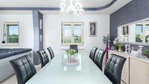 A modern dining room with a glass table, black leather chairs, a chandelier, and a view of greenery outside. - Villa Sv. Anna by Villas Guide (Radetici)