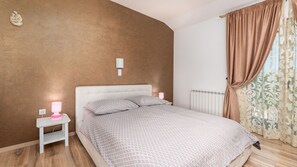 A bedroom with a white bed, patterned bedding, a nightstand with a lamp, a radiator, and a window with curtains. - Villa Sv. Anna by Villas Guide (Radetici)