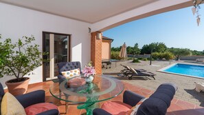 A glass-top table with chairs and a potted plant on a patio with a pool and outdoor furniture.