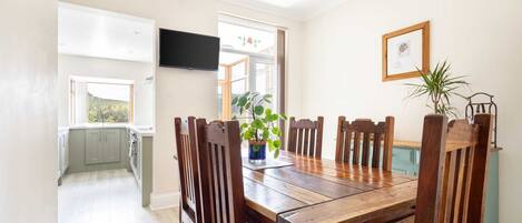 Uma área de refeições com mesa e cadeiras de madeira, uma planta em vaso e uma televisão fixada na parede.