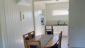 Uma área de refeições com uma mesa e cadeiras de madeira, uma cozinha ao fundo e uma janela com persianas.