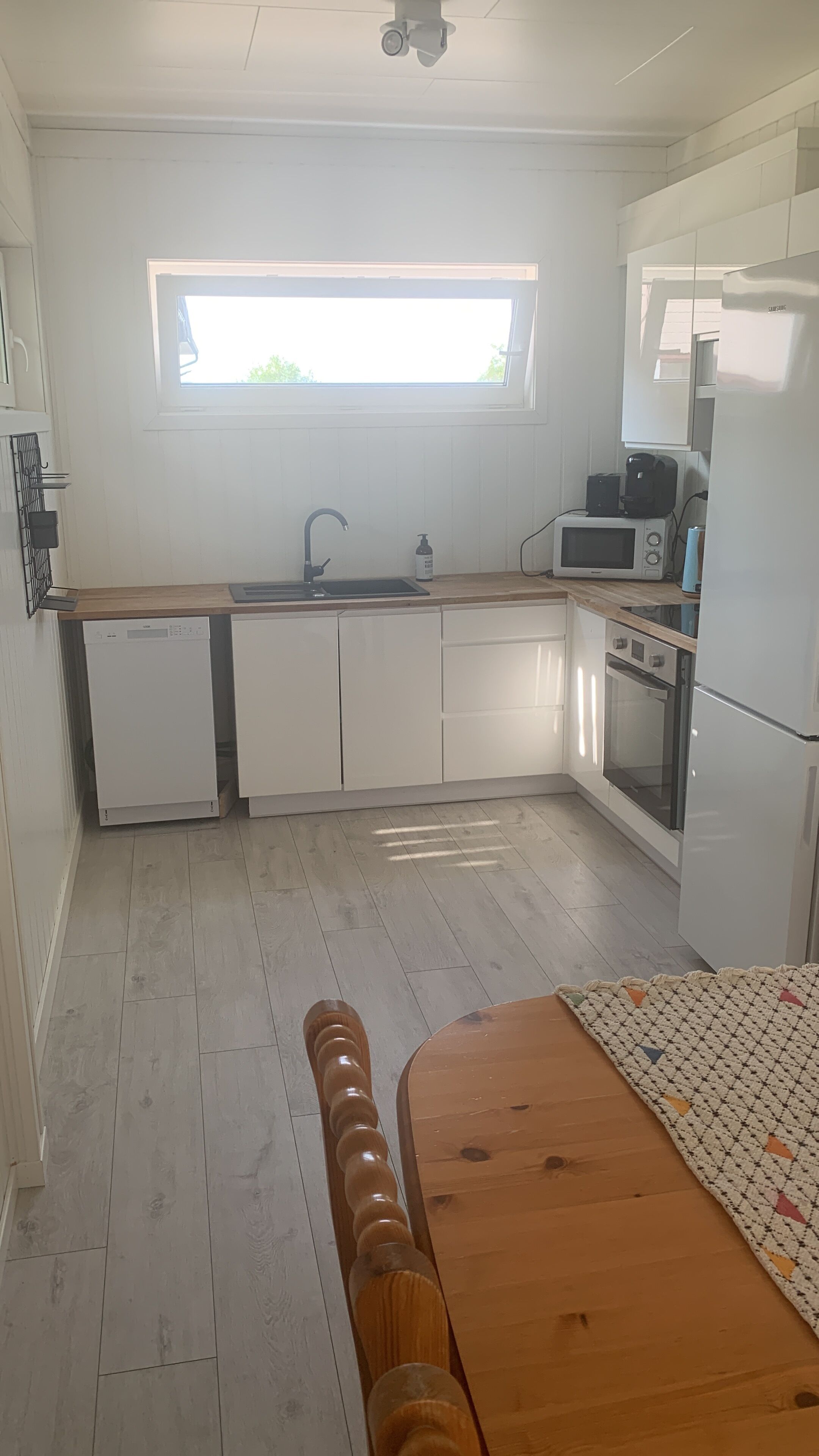Eine kompakte Küche mit Fenster, weißen Schränken, einer Spülmaschine, einer Mikrowelle und einem Kühlschrank.