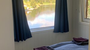 Quarto com uma grande janela com vista para um lago e montanhas, uma cama com roupa de cama listrada e piso de madeira.