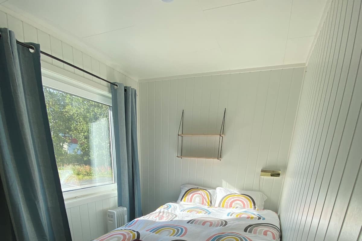 Une chambre avec un lit, une fenêtre avec des rideaux, un radiateur et une petite étagère.