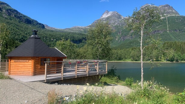 Uma cabana de madeira com varanda e vista para um lago, rodeada por montanhas.