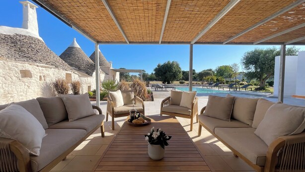 A covered outdoor seating area with two sofas and a coffee table, overlooking a pool and a stone building. - A Trullo Like No Other - Timeless Style, Modern Comforts. Private Pool (Selva di Fasano)