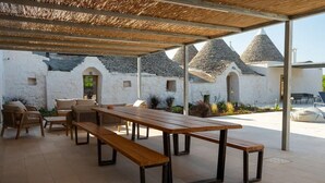 An outdoor patio with wooden benches and chairs, a thatched roof, and stone buildings in the background. - A Trullo Like No Other - Timeless Style, Modern Comforts. Private Pool (Selva di Fasano)