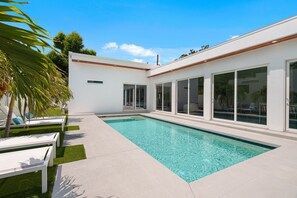 Ett modernt poolområde med ett kristallklart pool, vita solstolar och en vit byggnad med stora fönster.
