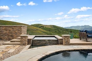 Une piscine en pierre agrémentée d’une cascade, entourée d’une terrasse avec des sièges et offrant une vue sur des collines et des montagnes ondulantes.