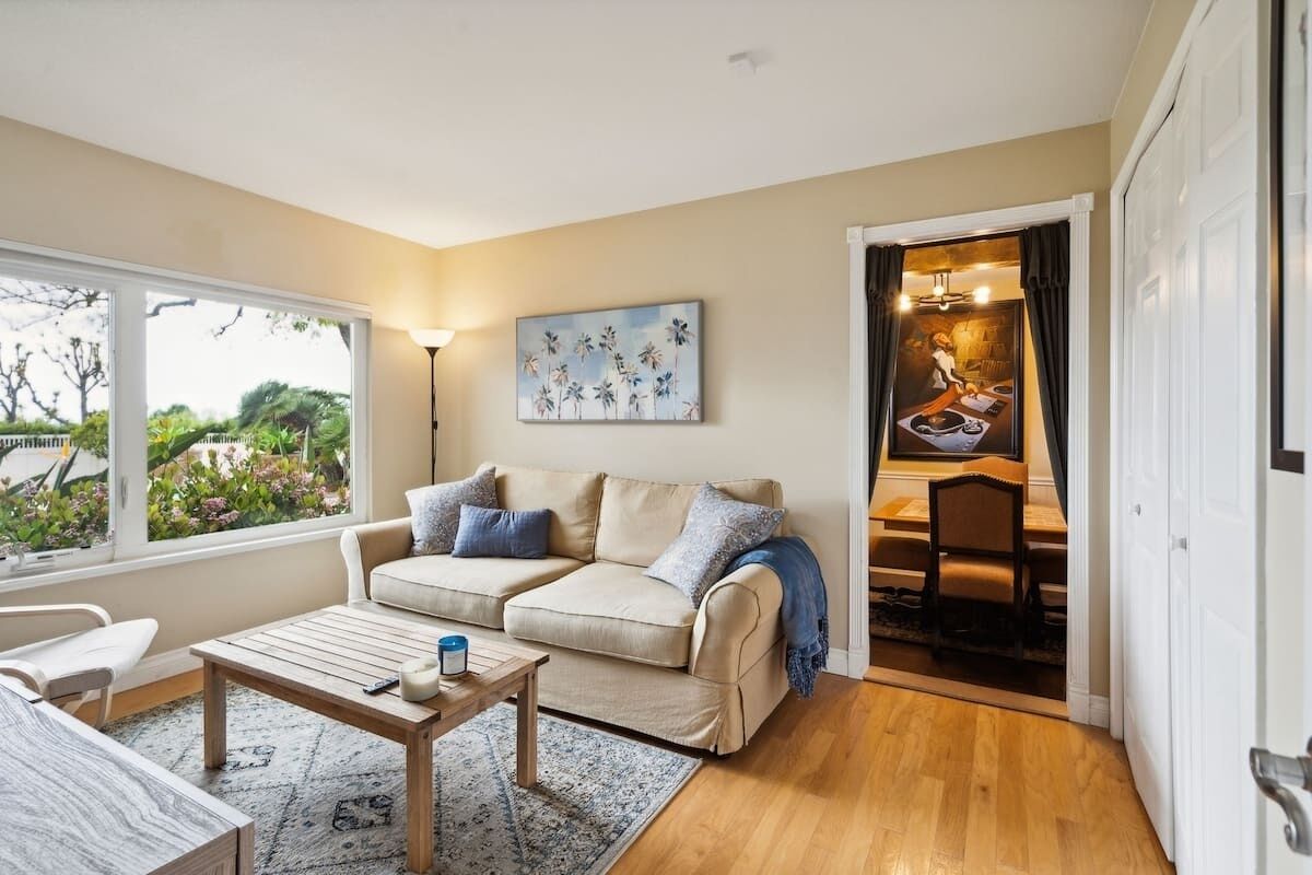 Un salon chaleureux avec un canapé beige, une table basse en bois et une vue sur un jardin, visible à travers de grandes fenêtres.