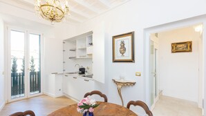 A dining area with a wooden table, chairs, a vase with flowers, a chandelier, a kitchen area with white cabinets, and a balcony with a view. - Amazing Domus Ruspoli  (Roma)