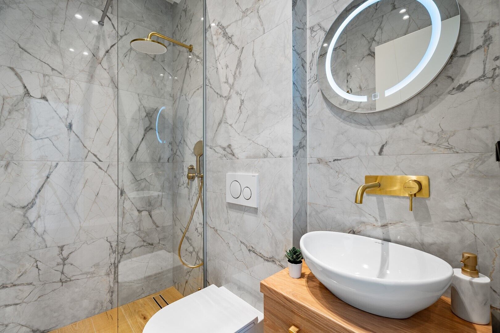 A modern bathroom with a marble shower, a round mirror, and a white sink with gold fixtures.