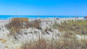 A beach with clear skies, sandy dunes, and a few people in the distance.