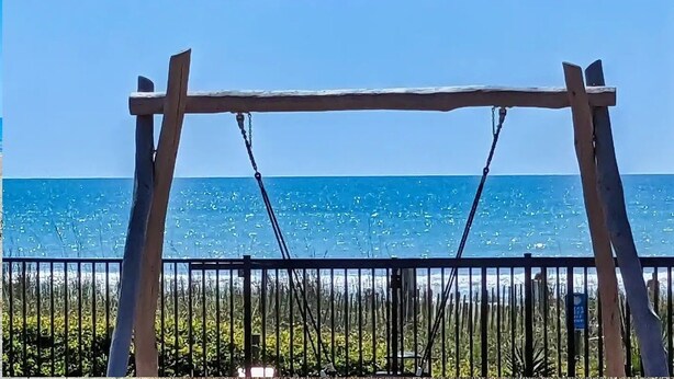 A wooden swing set with a metal fence in the foreground and a clear blue sky with the ocean in the background.