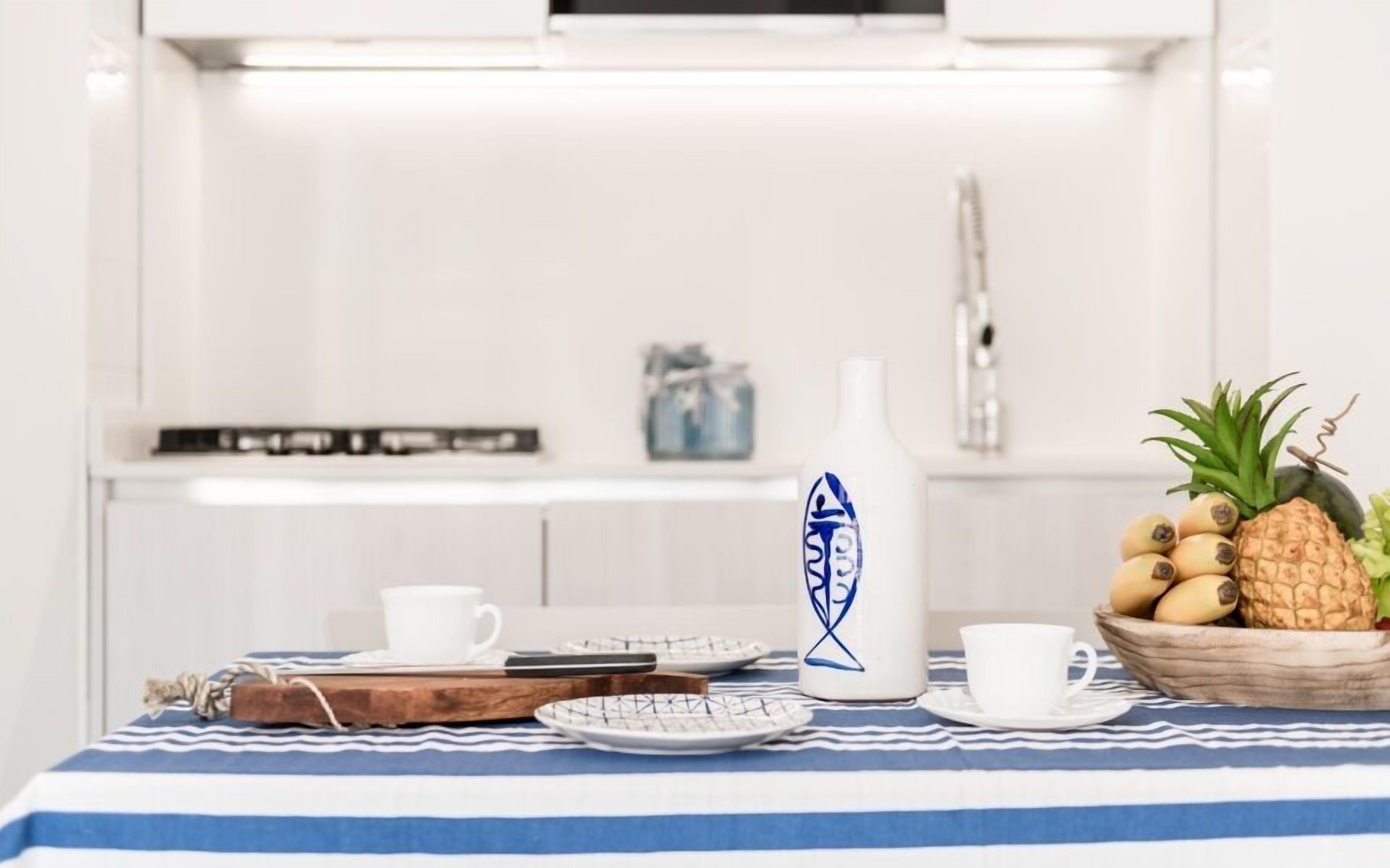 A kitchen with a striped tablecloth, a coffee pot, a bowl of fruit, and a white mug.