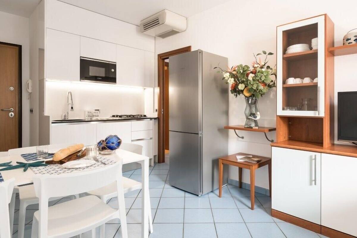 Una cocina moderna con armarios blancos, un refrigerador de acero inoxidable, un microondas y una pequeña mesa de madera con un florero.