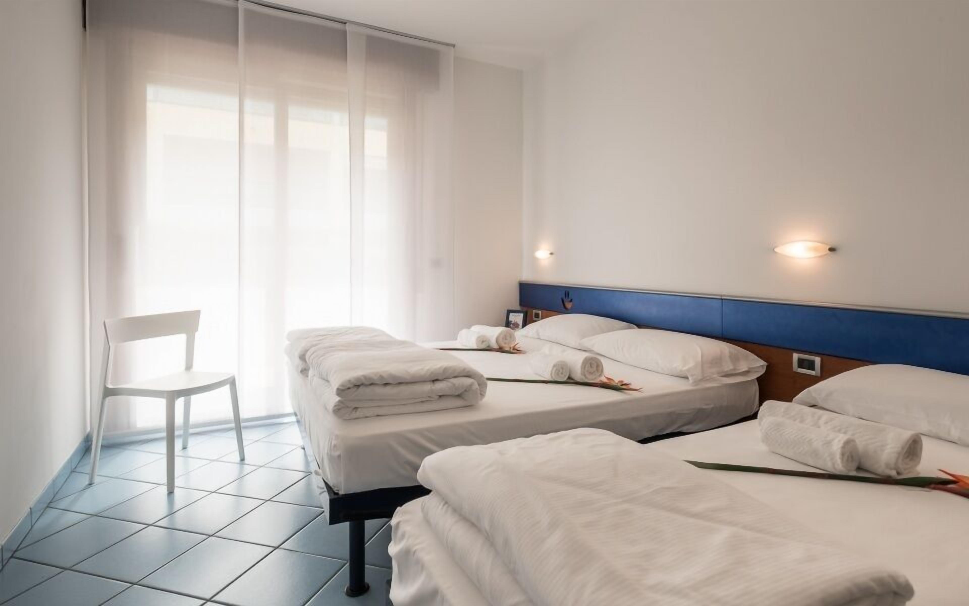 Une chambre d’hôtel avec deux lits, une chaise et une grande fenêtre avec des rideaux légers.