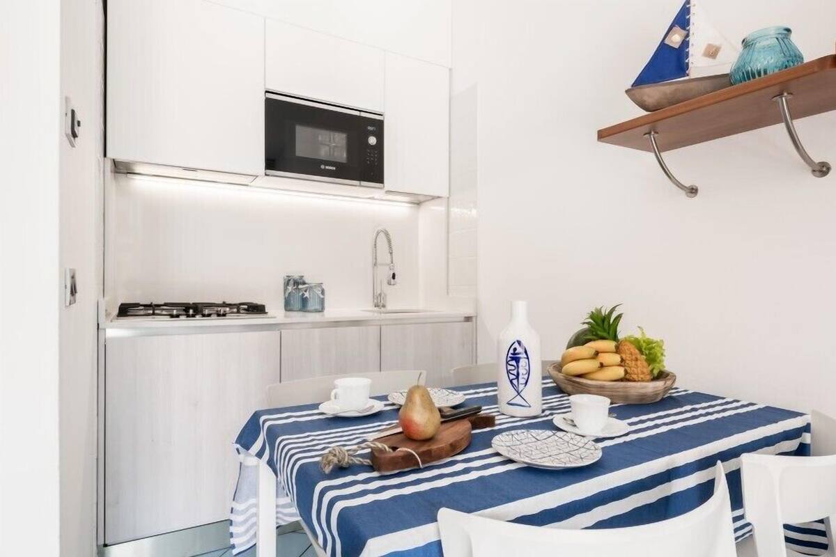 Una cocina compacta con armarios blancos, un microondas y una pequeña estufa a gas. Una mesa de comedor con mantel a rayas y un frutero está preparada para dos comensales.