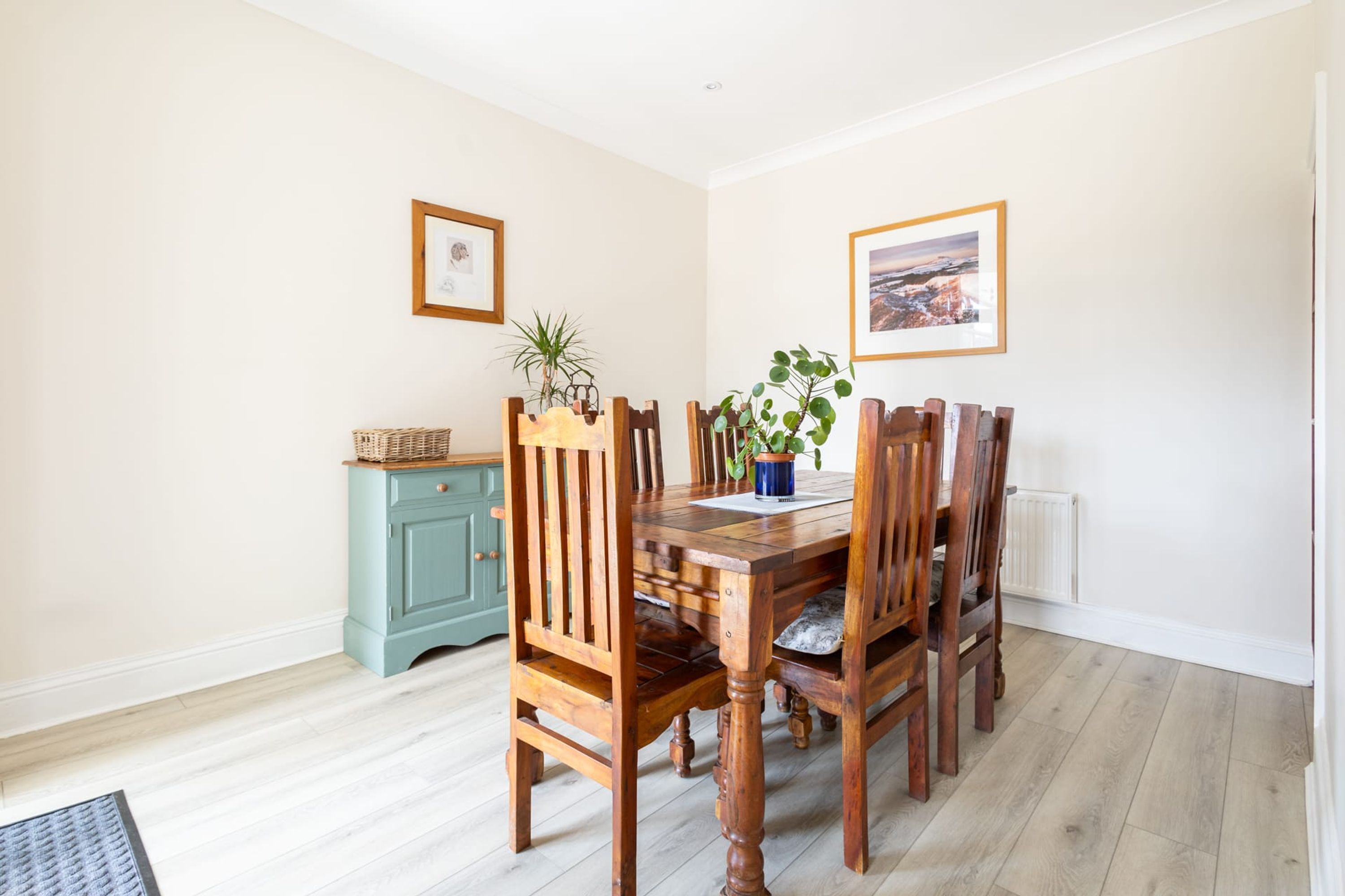 A dining area with a wooden table and chairs, a small cabinet, and two framed pictures on the walls.