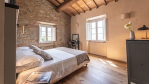 A bedroom with a stone wall, wooden ceiling beams, a bed with white linens, and two windows. - Typical Stone House with Swimming Pool 6/8 Pax in the countryside between Florence and Lucca. (marliana)