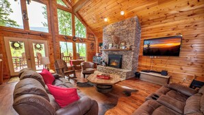 A cozy cabin interior with a stone fireplace, leather sofas, and a large window overlooking the outdoors. - *Gorgeous 3BD Cabin w/ VIEWS! Hot Tub + Fire Pit!* (Sevierville)