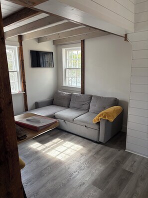 Una habitación con sofá gris, vigas de madera, paredes blancas y una ventana.