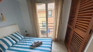 A hotel room with a bed, a small table with a lamp, a window with curtains, and a wooden door with louvered panels.