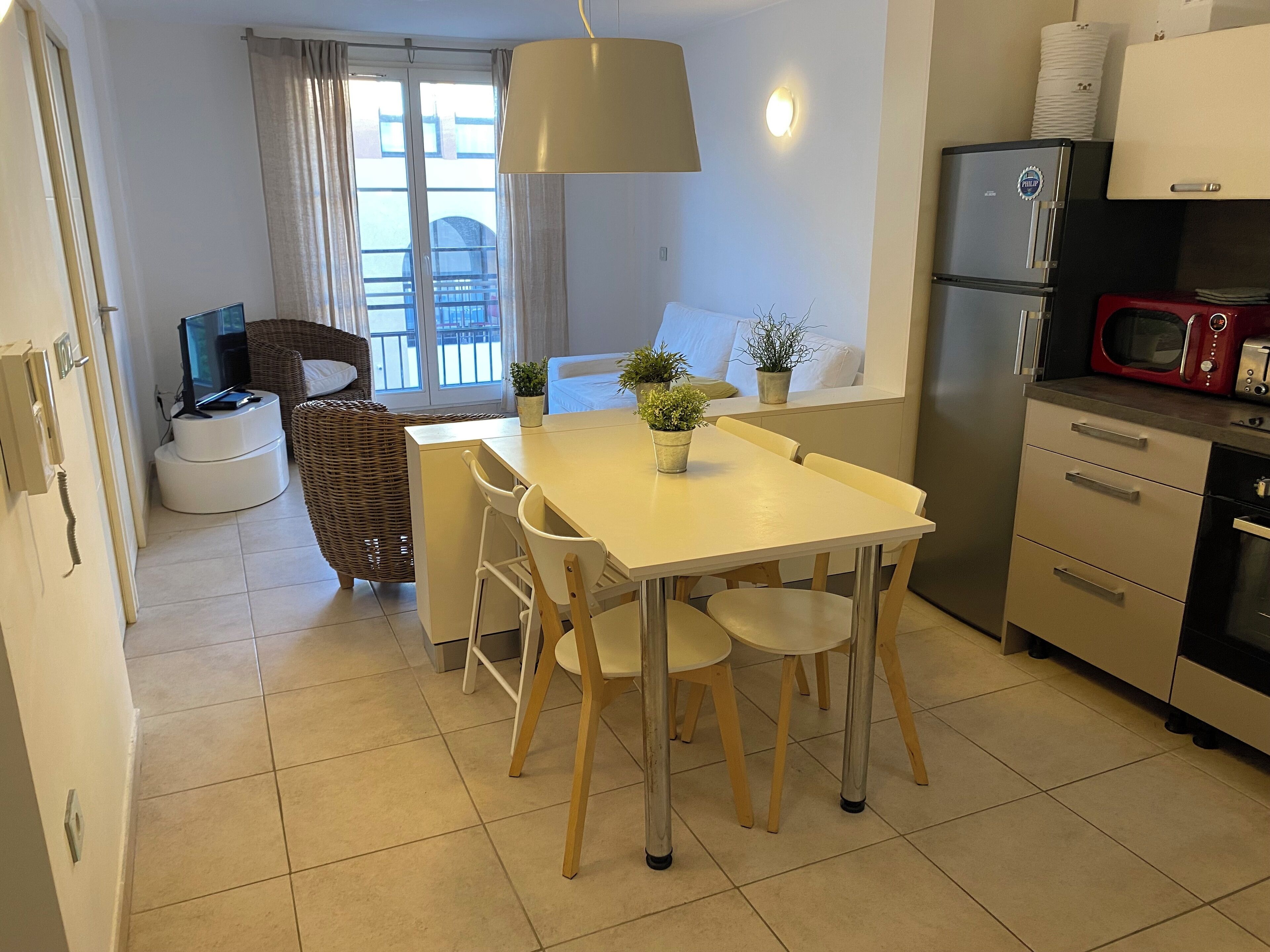 Una cocina compacta con una mesa de comedor, sillas, un refrigerador, un microondas y una pequeña zona de comedor con una silla de mimbre y un televisor.