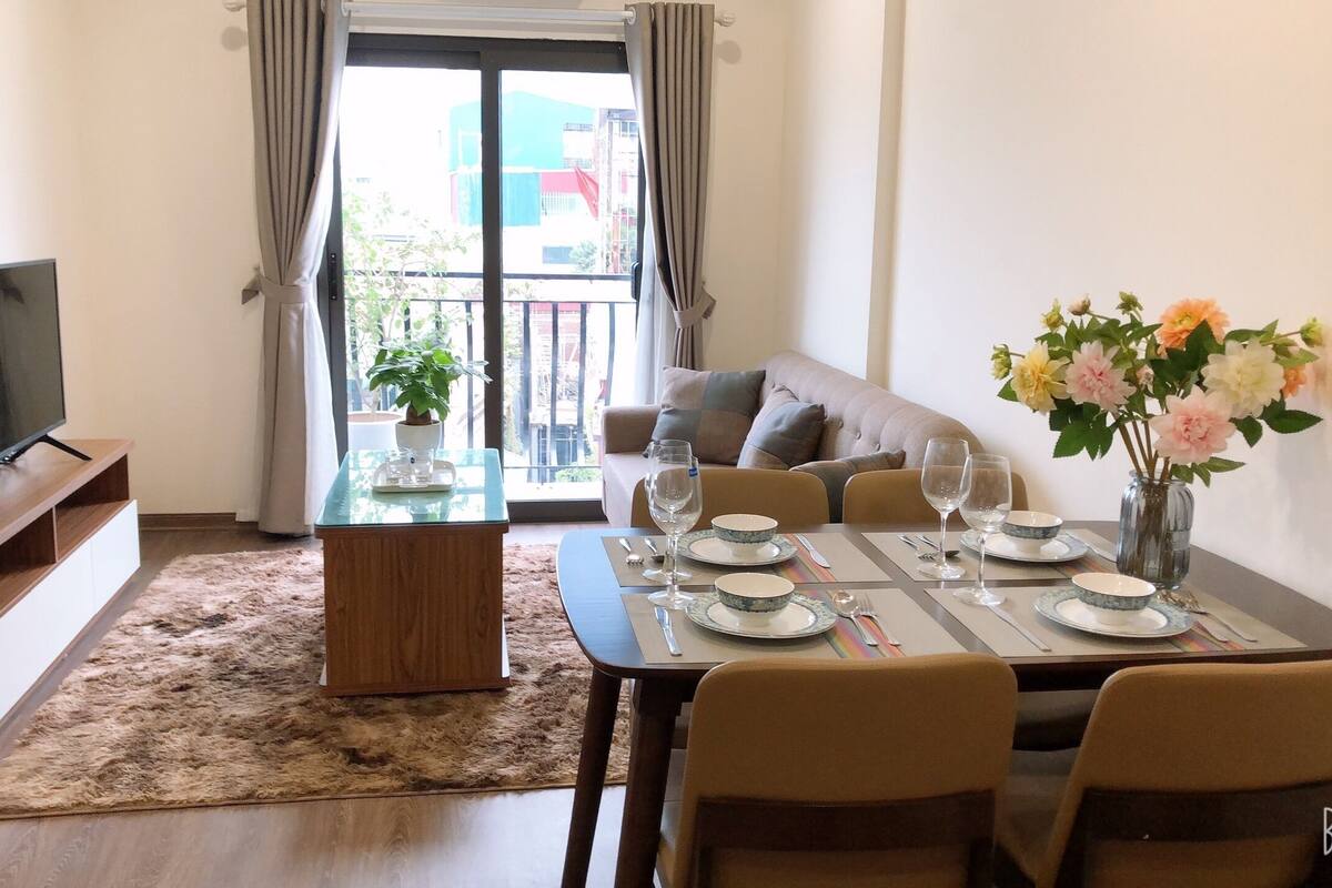 A dining area with a table set for four, a sofa, a TV, and a balcony with a view.