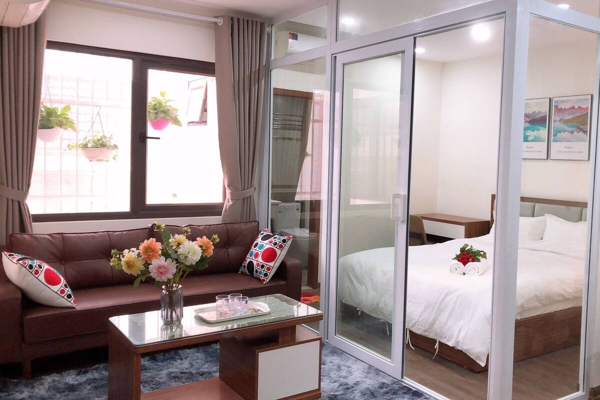 A hotel room with a sofa, a glass coffee table, a bed with a white duvet, a desk with a chair, and a sliding glass door leading to a balcony with plants.