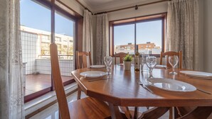Un comedor con una mesa de madera dispuesta para cuatro personas, ventanas amplias con cortinas y vistas a edificios.