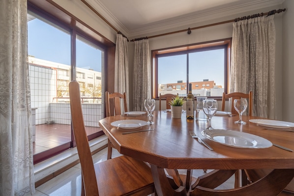 A dining area with a wooden table set for four, large windows with curtains, and a view of buildings outside.