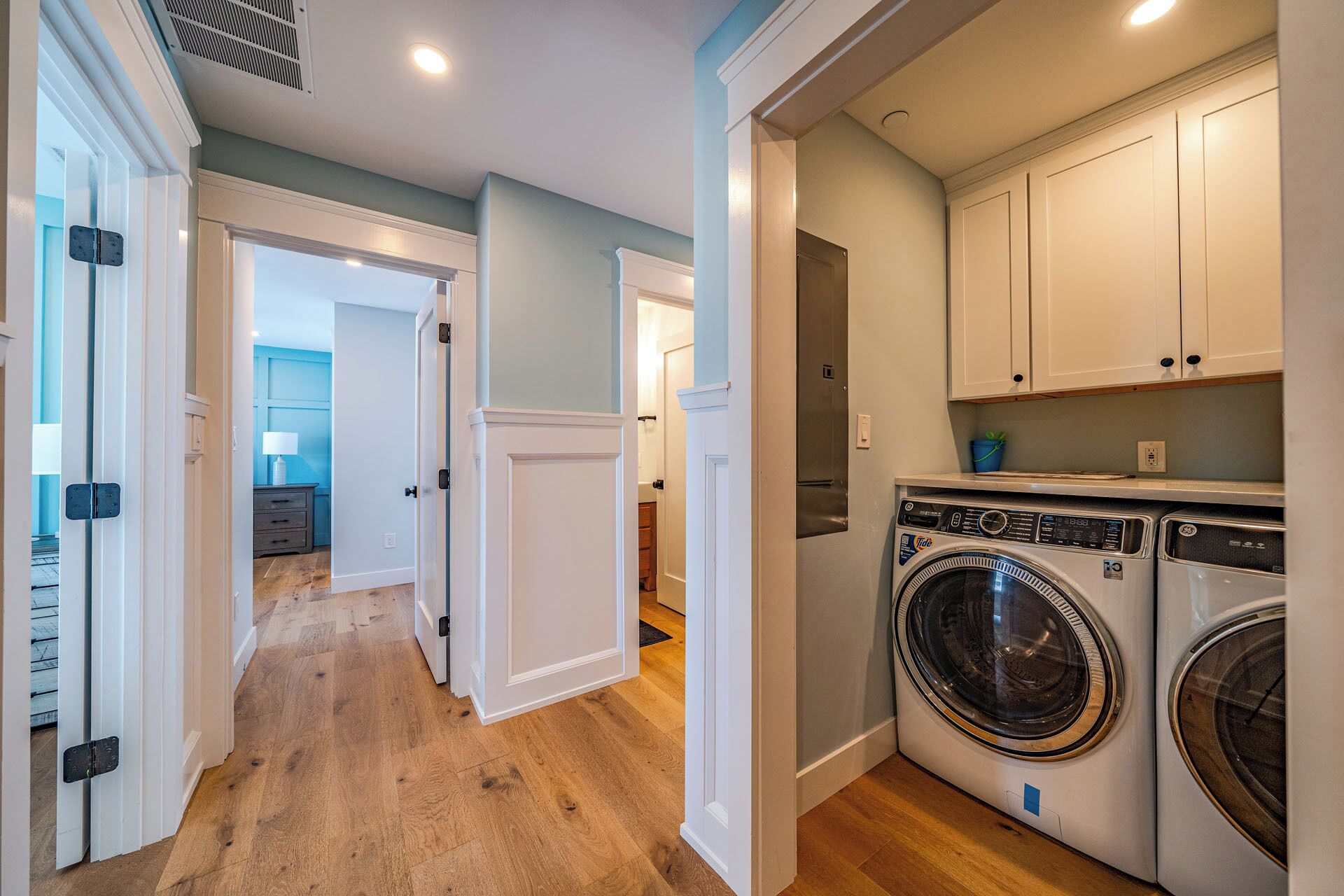 A laundry room with a front-loading washer and dryer, wooden flooring, and white cabinetry.