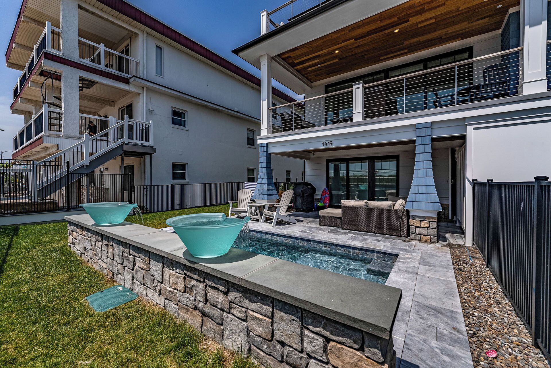 Une maison moderne à deux étages, dotée d’un mur de soutènement en pierre, d’une piscine, de mobilier d’extérieur et d’un patio couvert.
