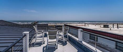 A rooftop terrace with a view of the beach, featuring outdoor seating and a railing.