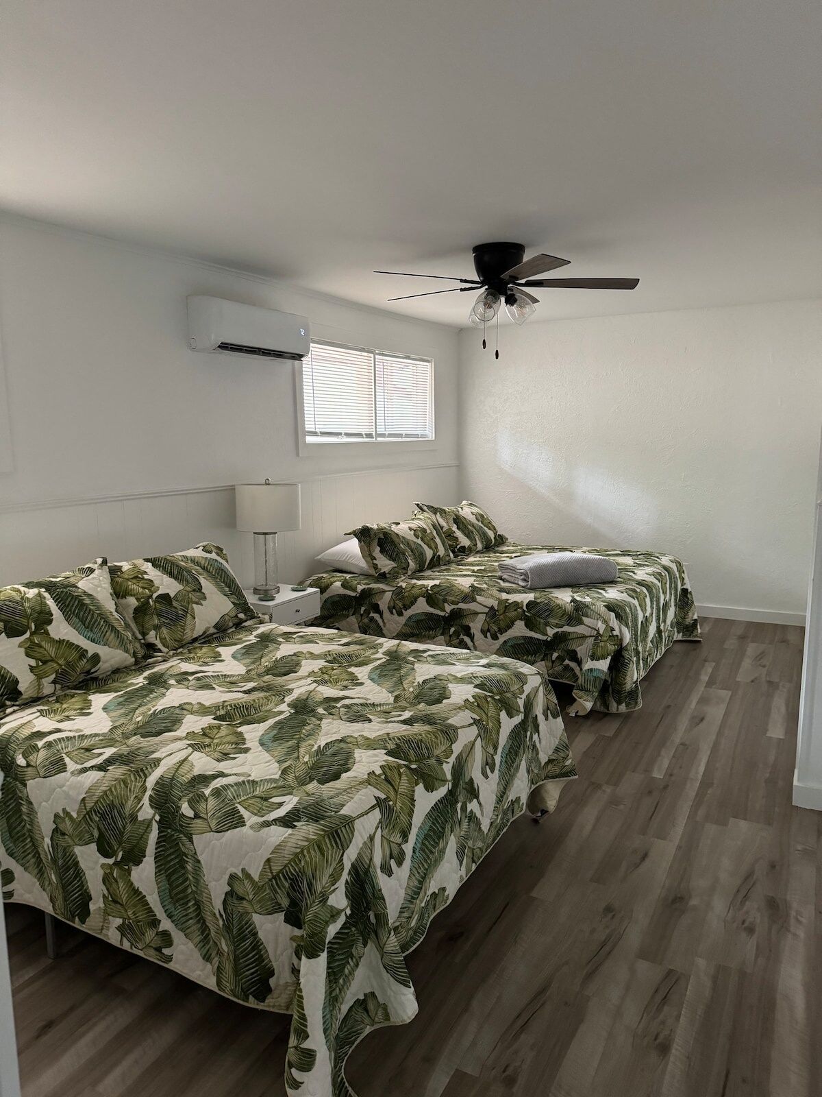 A bedroom with two beds, a ceiling fan, an air conditioner, and a window with blinds.