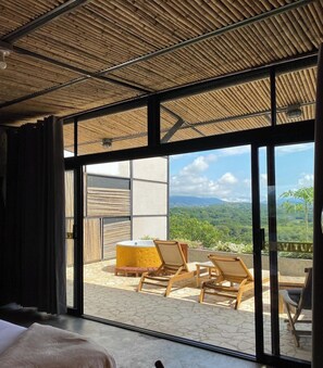 Een kamer met een grote schuifglazen deur die uitkomt op een terras met houten stoelen en een gele tafel.
