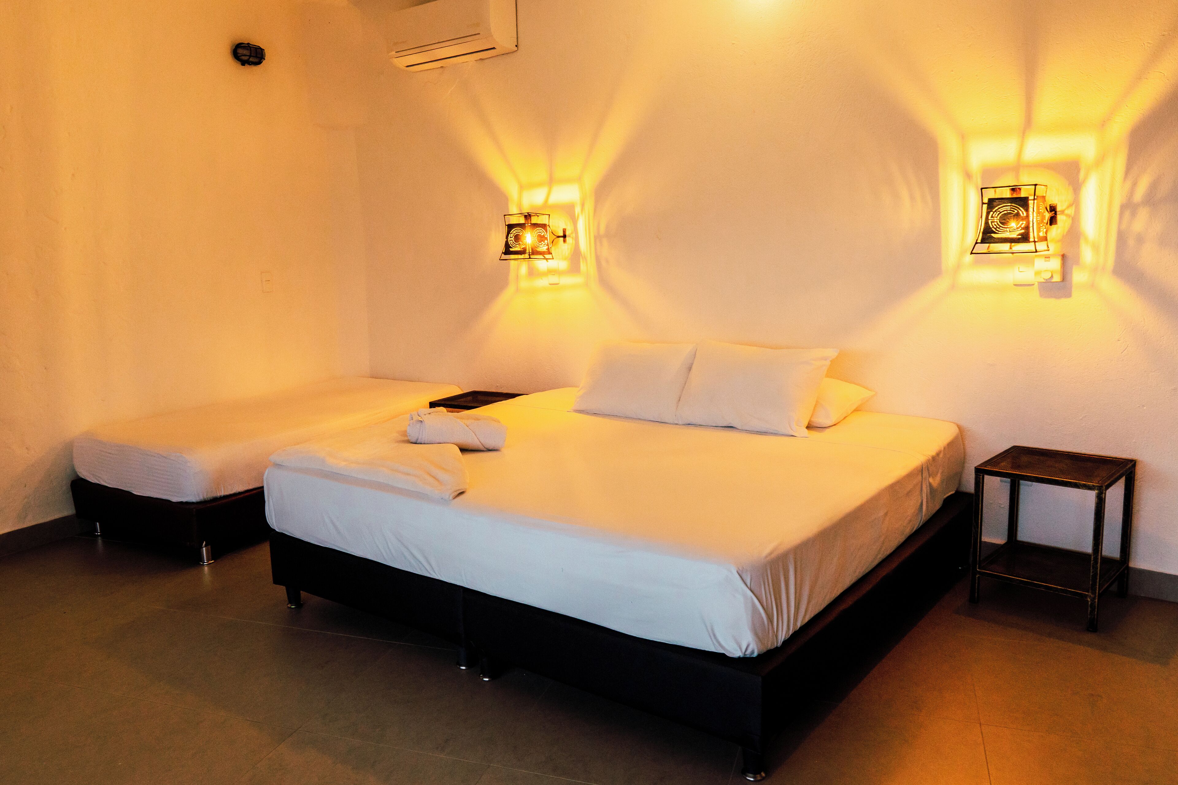 Una habitación de hotel con dos camas, una mesita de noche pequeña e iluminación instalada en la pared.
