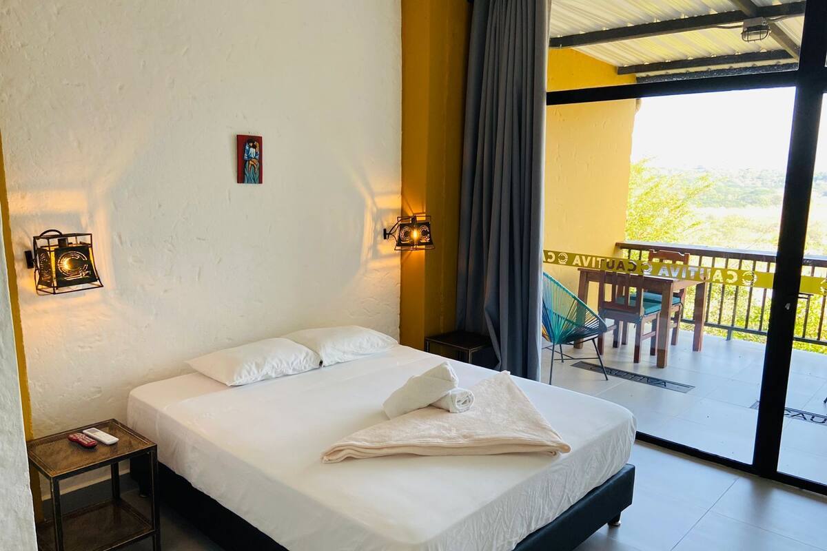 A hotel room with a bed, two bedside tables, a nightstand, a window with curtains, and a balcony with a table and chairs.