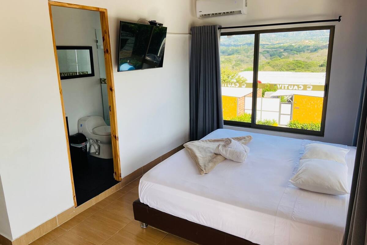 A hotel room with a bed, a TV mounted on the wall, an air conditioning unit, and a window with a view of the outside.