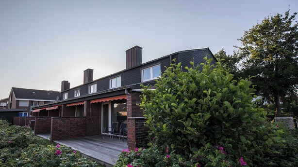 A residential house with a brick exterior, a wooden deck, and a garden with pink flowers.