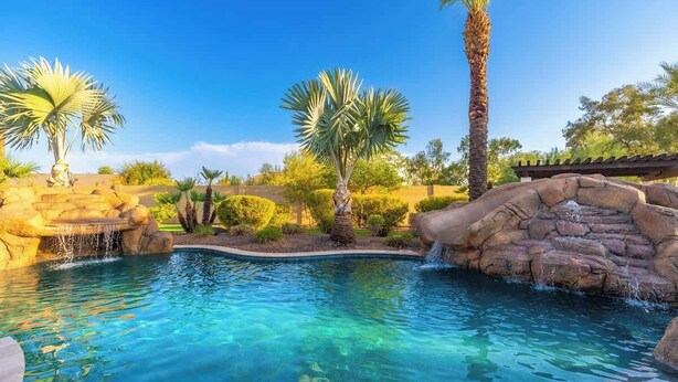 Una piscina cristallina con cascata, circondata da palme e una formazione rocciosa.