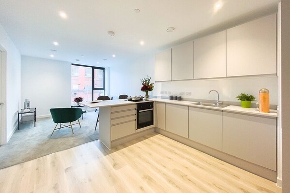 A modern kitchen with white cabinets, a black oven, and a sink. There is a dining area with a green chair and a small table. - Manchester Terrace Retreat-cinema & Free Parking (Manchester)
