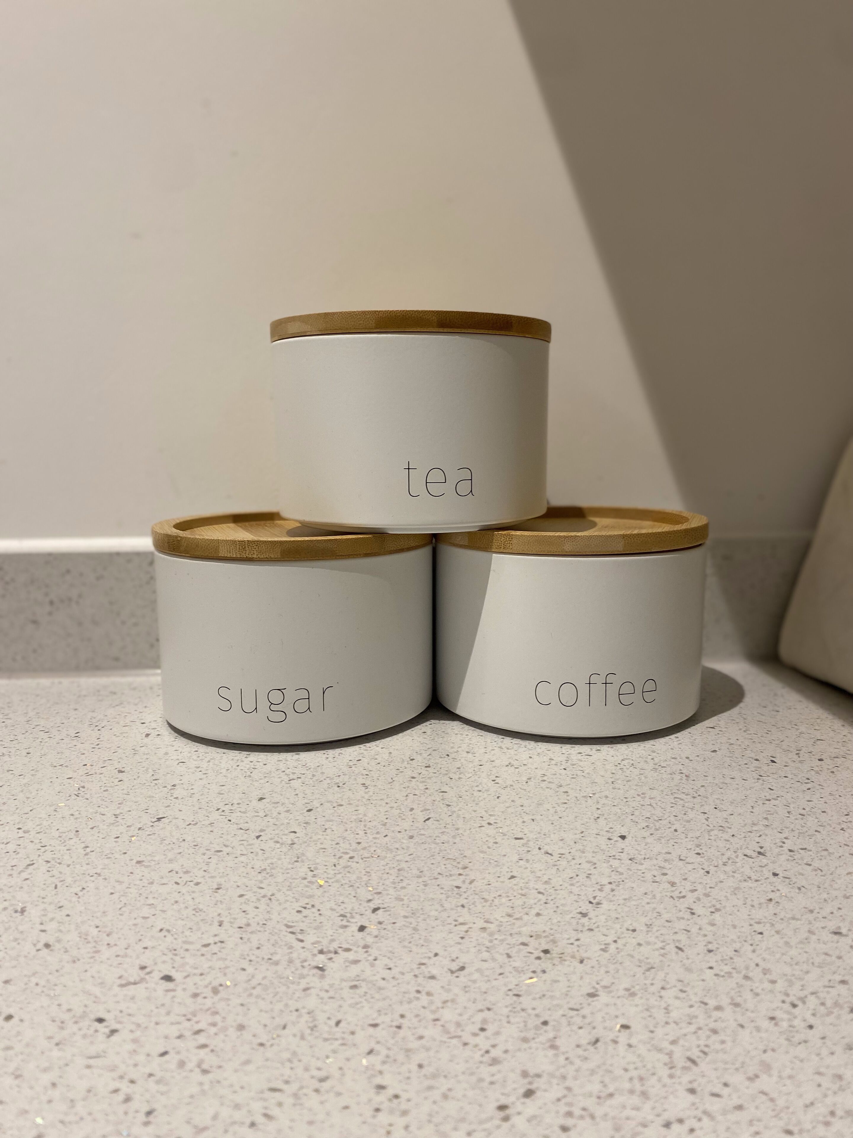 Três recipientes com os dizeres 'chá', 'açúcar' e 'café' em uma bancada estampada.