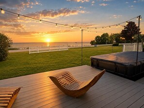 A deck with a hammock, a bench, and a hot tub, overlooking a grassy area and the ocean at sunset. - Castleview Cove- 5Bd 4Ba Lakefront Luxury Retreat! (Port Sanilac)