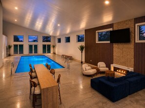 Ein moderner Poolbereich im Innenbereich mit einem langen Holztisch und Stühlen, einem blauen Sofa und einem Kamin.